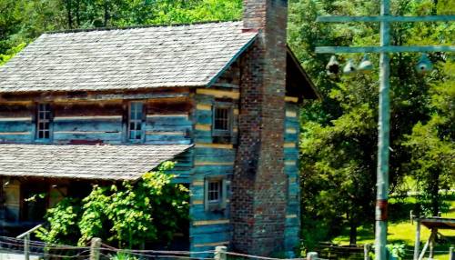 Historic log cabin with a stone chimney at the Clinton Museum in Tennessee, surrounded by greenery and wooden fencing.