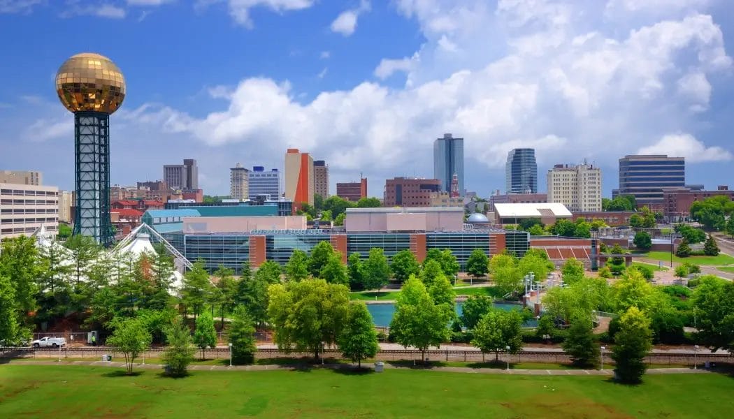 Skyline of downtown Knoxville, Tennessee with the Sunsphere and city buildings surrounded by green trees under a partly cloudy sky